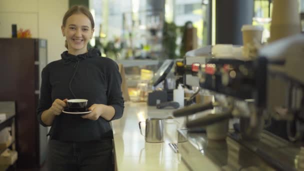 Wanita cantik muda tersenyum di depan kamera dan meregangkan cangkir dengan kopi segar yang lezat. Potret barista perempuan Kaukasia profesional di tempat kerja di kantin atau restoran. — Stok Video