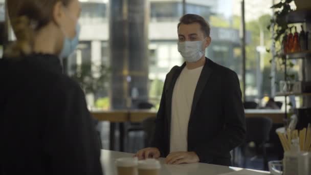 Pozitivní mladý podnikatel v Covid-19 obličejové masky dostávají kávu s sebou v kavárně a odcházejí. Portrét spokojeného zákazníka kupujícího osvěžující nápoj v dopoledních hodinách na pandemii koronaviru. — Stock video