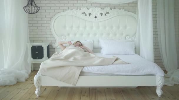 Wide shot van jonge mooie vrouw wakker in de ochtend. Gelukkig ontspannen Kaukasische dame liggend in groot wit bed in de slaapkamer en stretching. Weekend vrijetijdsconcept. — Stockvideo