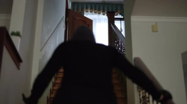 Siyah giyinmiş ve maske takmış bir kız soygun sırasında elini duvara dayayıp üst kata çıkıyor..