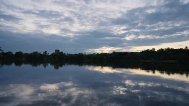 Bulutlu dramatik gökyüzü su yüzeyine yansıyan huzurlu bir akşam gölü manzarası. Ufuktaki ağaçlar sakin sahneyi çerçevelerken hafif dalgalanmalar doku oluşturur..