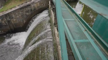 Güçlü su akıntıları, bakım erişimi için kenar boyunca uzanan yeşil metal bir geçitle küçük bir hidroelektrik barajının beton sızıntısından aşağı taşar..