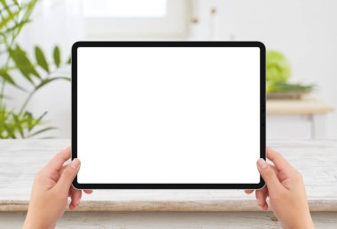 İzole edilmiş insan sol elinde siyah tablet medya aygıtı beyaz ekran modeli ve mutfakta ahşap masa var.