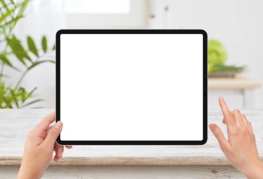 İzole edilmiş insan sol elinde siyah tablet medya aygıtı beyaz ekran modeli ve mutfakta ahşap masa var.
