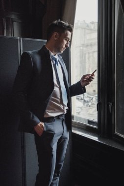 Ofiste bir pencere yanında duran genç bir işadamı yan görünüm. Onun akıllı telefon beklerken kontrol ediyor.