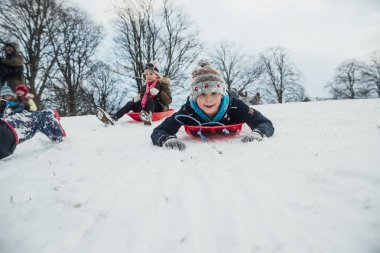 Küçük bir çocuk arkadaşlarıyla bir kızağa karlı tepeden geliyor olarak kameraya bakarak düşük açılı görünüş.