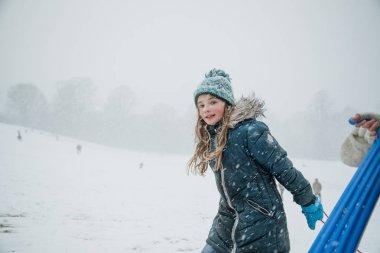 Küçük kız yokuş yukarı bir kızak yarışı için arkadaşları ile başlık ise kameraya bakıyor.