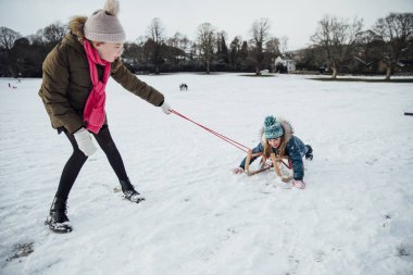 İki küçük kız karda eğleniyorlar. Bir kız diğer bir kızak üzerinde çekiyor.