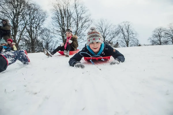 Küçük bir çocuk arkadaşlarıyla bir kızağa karlı tepeden geliyor olarak kameraya bakarak düşük açılı görünüş.
