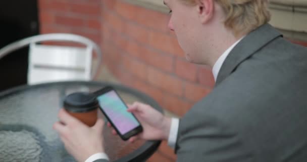 在一个年轻的商人坐在一张桌子的肩膀视图 用他的智能手机喝一杯带走的咖啡 — 图库视频影像