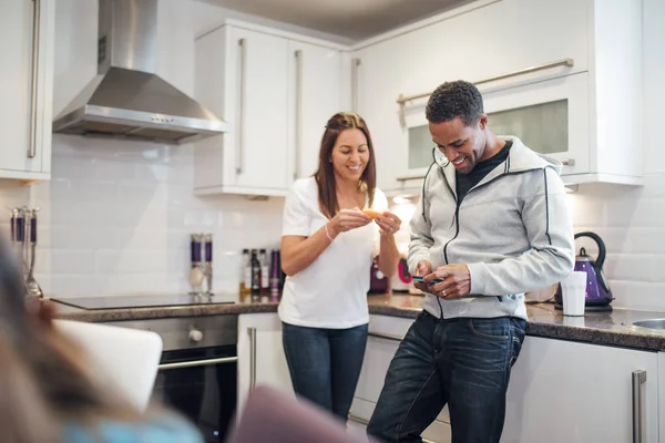 Mid adult couple are relaxing in the kitchen of their home together. The woman is eating a slice of orange and the man is using his smart phone.