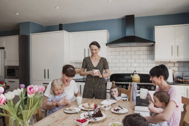 Üç yetişkin kadınların kızları ile mutfakta orta. Kızlarının onların öğle yemeği yerken kucaklarında üzerinde oturan iki kadın oturuyorsun. Bir yetişkin kadın orta bir bardak çay ile ayakta ve gülümseyen.