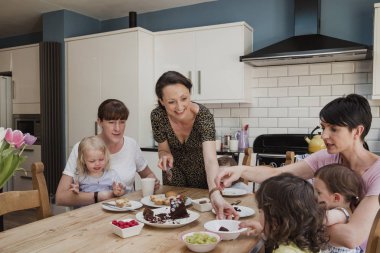Yetişkin kadın bir dilim pasta küçük bir kız için teslim yemek masasında oturdu. Orada iki yetişkin kadın çocukları tatlı davranıyor zevk ile masaya oturdu orta diğer.