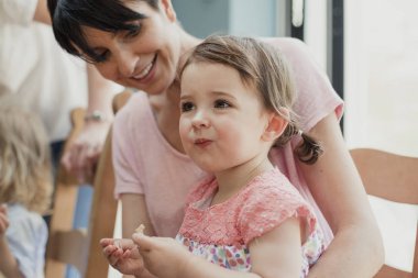 Yan görünüm oturan küçük bir kız onu annelerin kucak. O yeni bir gıda çalışıyor ve o çalışırken bir yüz çekerek. Annesi gülümseyerek ve ona bakarken gülüyor.