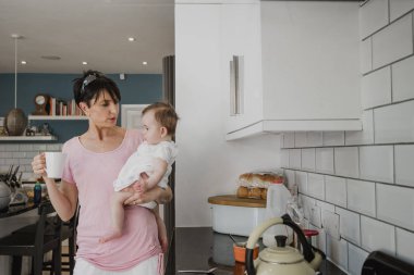 Mutfakta bebek kızı dururken tutan bir anne önden görünümü. Kadın onun tarafında kızı taşırken bir fincan çay tutuyor.