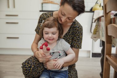 Önden Görünümü bazı ahududu ile oynayan ve parmaklarını koyarak küçük bir kız. Annesi ona yardım arkasında diz çökmüş ve onunla gülüyor.
