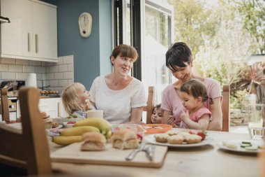 İki yetişkin kadın mutfakta yemek masası ile iki kızı kucaklarında üzerinde oturan orta. Anneleri konuşmak ve gülümseme iki küçük kız onların öğle yemeği tadını çıkarıyor.