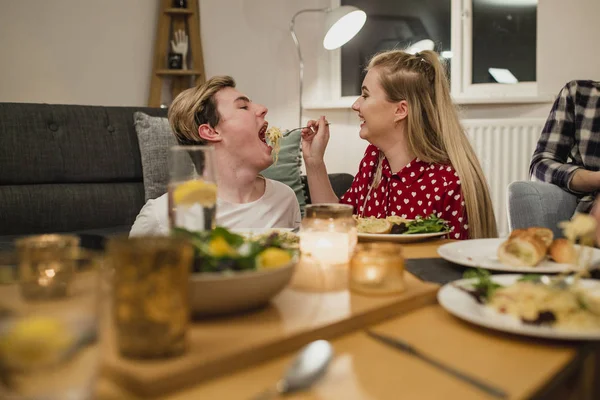 Young couple are at a dinner party and the girl is feeding her partner a big mouthful of spaghetti carbonara.
