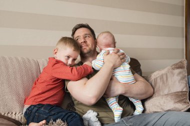 Baba ve iki oğlu kanepede oturan ve rahatlatıcı. Küçük çocuk, babasının sarılma ve tarassut belgili tanımlık tv. Babası onun yeni doğan bebek çocuk tutan ve onu geğirme.