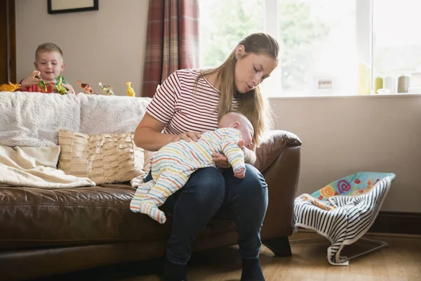 リビング ルームにはソファの端に座って 彼女の生まれたばかりの子供を彼を授乳後げっぷを母します 彼女の息子は彼のおもちゃながら再生するとソファの後ろに Stadning — ストック写真