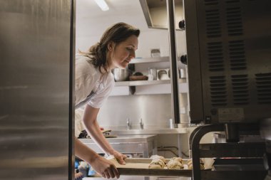Yetişkin kadın baker pişmemiş çörekler bir tepsi fırına yerleştirerek..