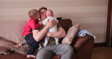 Baba ve iki oğlu oturma kanepe ve rahatlatıcı, küçük çocuk, babasının sarılma ve tarassut belgili tanımlık tv. Babası onun yeni doğan bebek çocuk tutarak ve onu sarma.