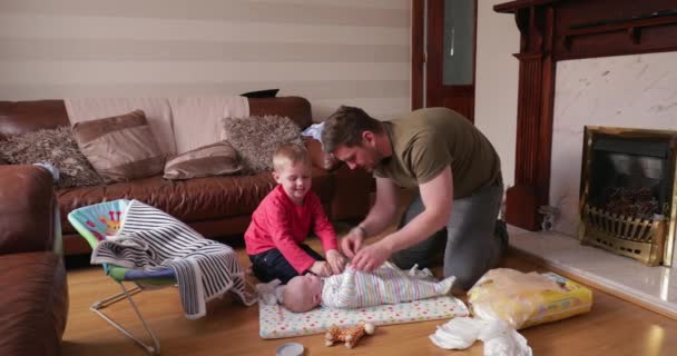 小男孩帮他爸爸在换尿布后把他的小弟弟扶起来 他们在客厅里给小男孩换尿布 在他的婴儿席上 — 图库视频影像