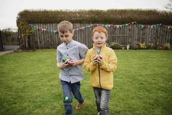 Two young boys walking through the back garden after an easter egg hunt. They are holding handfuls of chocolate eggs in their hands.