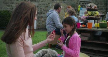 Küçük kız ve annesi dışarıda bir battaniyenin üzerinde oturmuş konuşurken. Küçük kız bir bahçe partisinde saçlarıyla konuşuyor ve oynuyor..