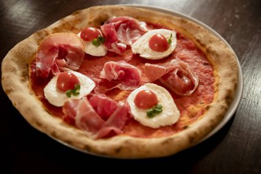 Domates, prosciutto ve mozzerlla pizzasının ahşap bir masadaki yakın çekimi..