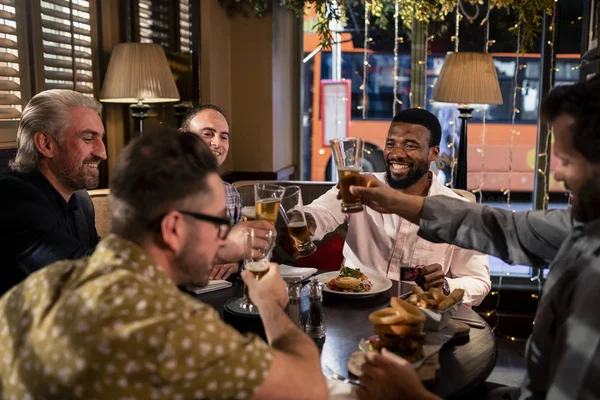 Small group of male friends sitting down in a restaurant and enjoying food and a beer together. They are making a Celebratory toast.