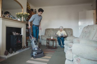 Genç çocuk, büyükannesi gazete okurken onun için oturma odasını süpürüyor.. 