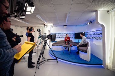 Stüdyoda çekilen bir TV şovu. Sunucular stüdyo kanepesinde oturup birbirleriyle konuşurken kamera ekibi onları film kameralarıyla çekiyor..