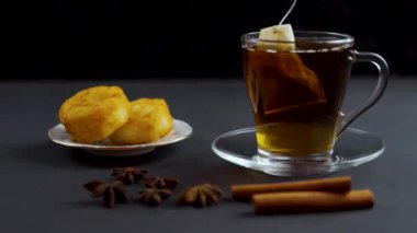 Çay poşeti siyah arka planda şeffaf bir bardakta demlenir. Peynirli kekler, anason ve tarçın bir fincan çayla siyah arka planda.