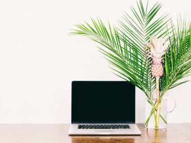 Laptop ile a boş siyah perde. Freelancer masaüstü whith bilgisayar, hurma dalları ve ahşap bir masa üzerinde bir vazoda dekoratif pembe ananas.