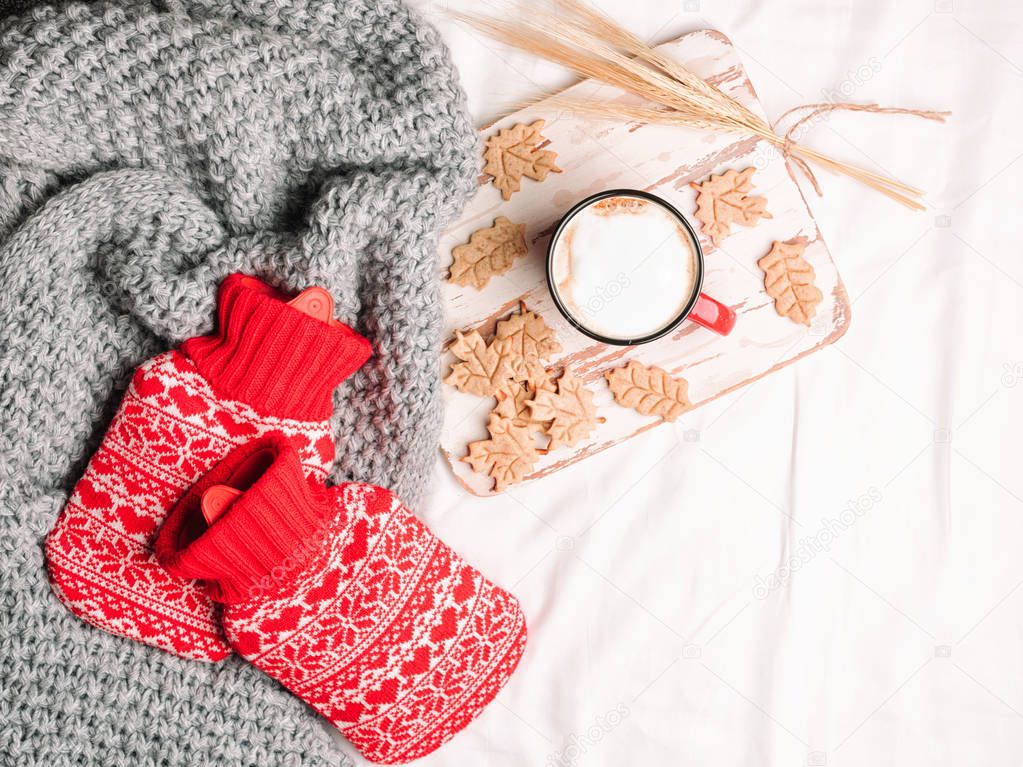 Red hot-water bottles on a bed. Heat pads on sheet, warm cozy grey knitted plaid and cup of coffee and cookies shape of a leaves on a tray. Top view or flat lay, copy space for your text
