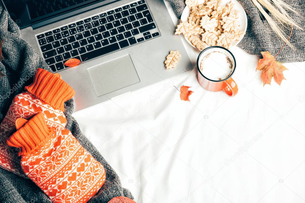 Workplace in a bed whith Cup of coffee and hot-water bottles. Hot drink on sheet, warm cozy grey knitted plaid, autumn leaves. Kraft envelope whith invitation and laptop. Top view or flat lay