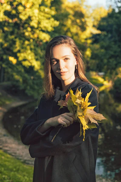 Portrait of young brunette woman in a black coat holds a bouquet of fall leaves. Autumn Weekend, windy warm sunny weather, fall colors