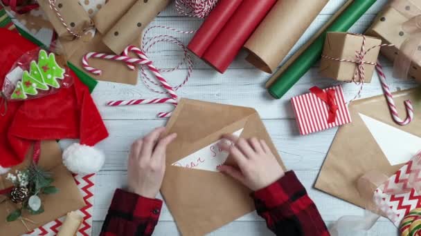 女性の十代の手ギフト木製テーブルにサンタさんへ手紙を書いて 赤のクリスマスの装飾をラップします クリスマスと新年あけましておめでとうございますの組成物 フラット横たわっていた トップ ビュー — ストック動画