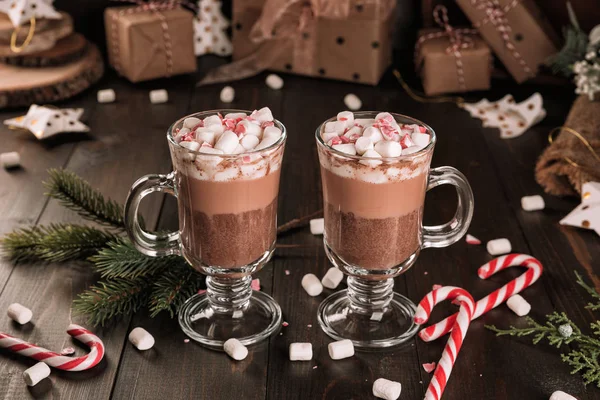 Cup of hot chocolate cocoa drink with a marshmallows and candy cane on wooden table. Two glasses of a sweet winter beverage and fir-tree branch.