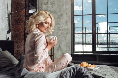 İpek Pembe pijama genç güzel kadın kahvaltı kahve bir kruvasan ile içme yatakta, var. Güneşli sabah, mavi gökyüzü harika görünümü penceresinden