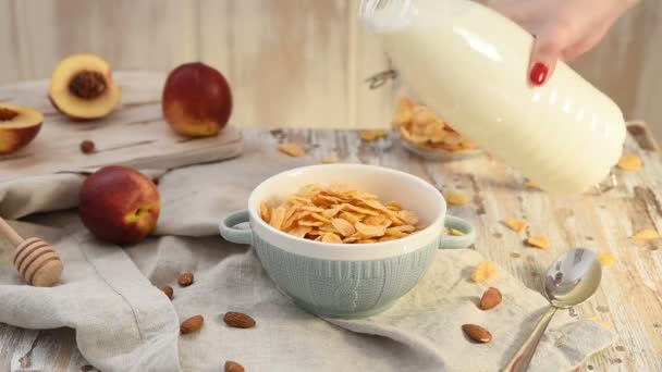 女人把牛奶从装在碗里的瓶子里倒入松脆的玉米片在木制桌子上 有杏仁和桃子的健康早餐 — 图库视频影像