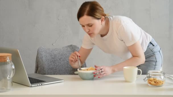 白いTシャツの若いブロンドの女性は ソーシャルメディアやラップトップ上のニュースフィードを見ている間 ミルクと高速朝食カリカリコーンフレークを食べる — ストック動画