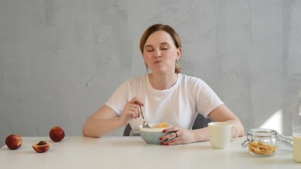 白いTシャツで疲れて眠い若いブロンドの女性はミルクと朝食のカリカリコーンフレークを食べる — ストック動画