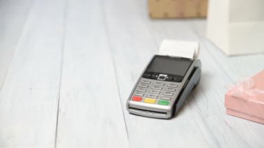 Kadın bir banka kartında NFC teknolojisi ile öder. Beyaz ahşap arka planda ödeme terminali. Hediye kutuları ve satış noktası terminali veya POS makbuzu ile. Metniniz için boşluk kopyalama