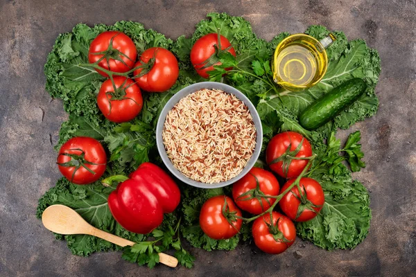 Kale salad leaf and red tomatoes, pepper, cucumbers with water drops on an old rustic metal tray. Green nature background. A bowl of brown rice, wooden spoon and olive oil