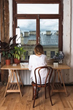 Beyaz bluzlu genç bir kadın ofis penceresinin yanındaki masada oturuyor. Dizüstü bilgisayarda çalışmak, kitap okumak, arka plan bakmak.
