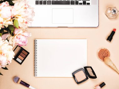 Dizüstü bilgisayarı, kozmetik ürünleri ve pastel pembe masasında şakayık çiçekleri olan bir blog yazarı çalışma alanı. Düz yatış, üst görünüm