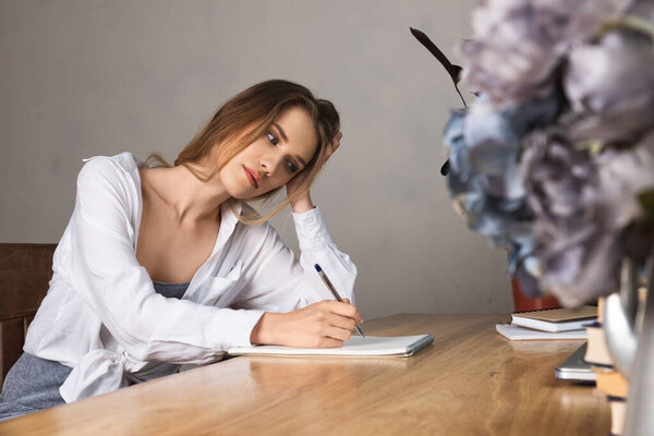 Dreaming young woman in a white blouse and grey dress sitting at a desk near the window in a home office. Writing or drawing