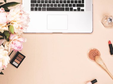 Dizüstü bilgisayarı, kozmetik ürünleri ve pastel pembe masasında şakayık çiçekleri olan bir blog yazarı çalışma alanı. Düz yatış, üst görünüm